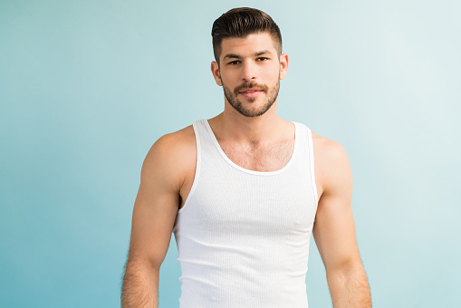 Confident attractive Hispanic male with blank expression wearing white vest isolated against turquoise background