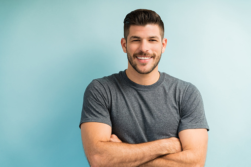 Portrait of handsome smiling Latin man standing with arms crossed against turquoise background