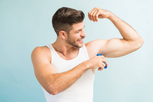 Attractive Hispanic Man Adding Fragrance To Muscular Body Smiling smart young Latin male applying antiperspirant in armpit while standing against turquoise background deodorant stock pictures, royalty-free photos & images