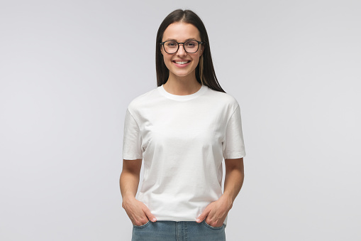 Young smiling woman standing with hands in pockets, wearing blank white tshirt with copy space, isolated on gray background