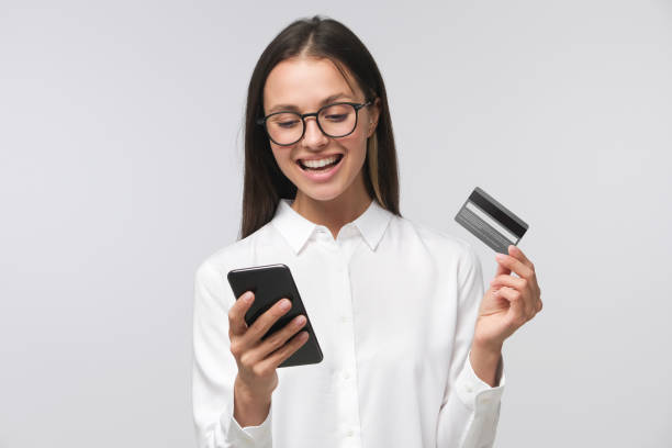 joven mujer sonriente sosteniendo tarjeta de crédito y teléfono inteligente, aislado en fondo gris - greeting card holding women credit card fotografías e imágenes de stock
