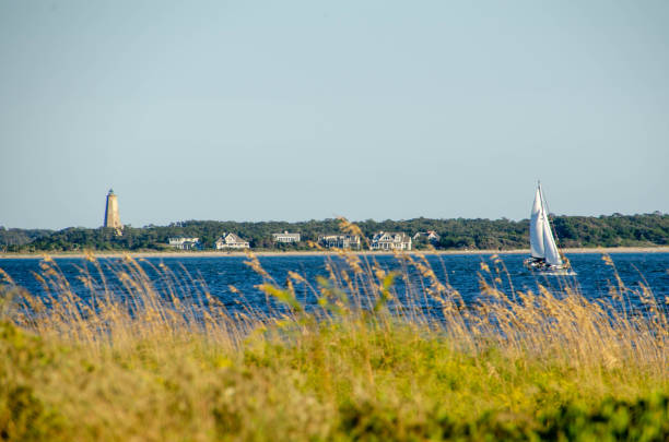Looking over the dunes and grass at a sailboat sailing past on the ocean. Looking past the dune grass a sailboat sails by. Baldhead Island is in the distance. cape fear stock pictures, royalty-free photos & images