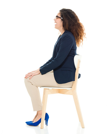 Full length / one person / side view / profile view of 30-39 years old adult beautiful curly hair / long hair latin american and hispanic ethnicity / puerto rican ethnicity female / young women businesswoman / business person sitting / resting in front of white background wearing eyeglasses / t-shirt / shirt / blazer - jacket / jacket / pants and high heels who is smiling / happy / cheerful / sitting on chair