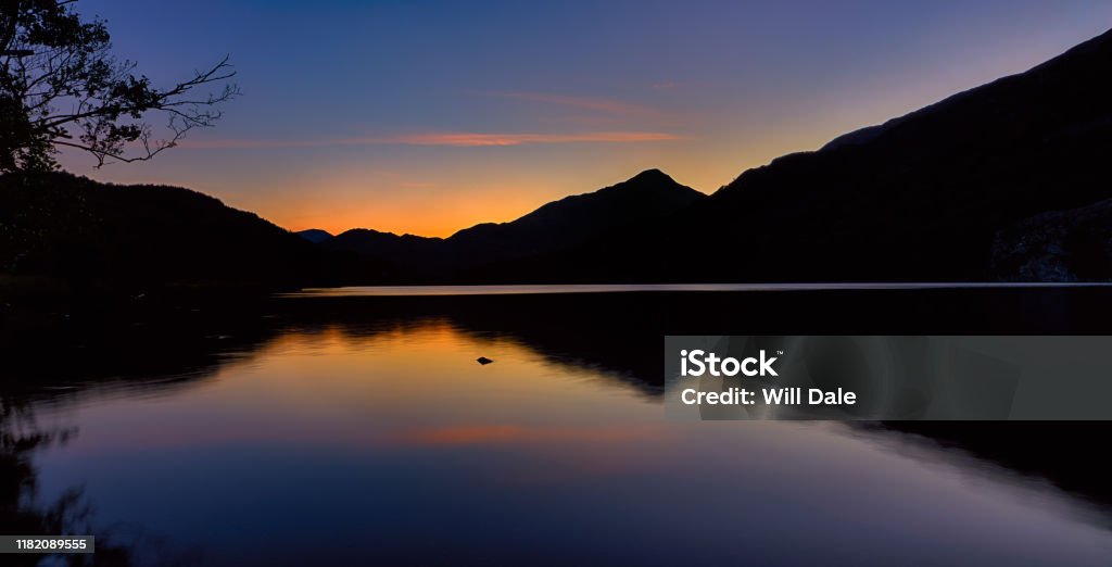 The sun has set behind the mountains around Llyn Gwynant in Snowdonia The sun has set behind the mountains around Llyn Gwynant, a lake in Snowdonia (Eryri), Wales (Cymru), UK. Silhouetting the mountains, casting a reflection off of the water. Llyn Gwynant Stock Photo