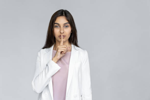 young adult indian businesswoman in white formal wear standing isolated on grey background with copy space. woman looking at camera and covering lips with finger, making silent sign, keep secret - silence finger on lips businesswoman one person imagens e fotografias de stock