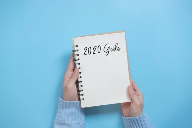 New Year 2020 goals list with notebook on blue background, flat lay style.