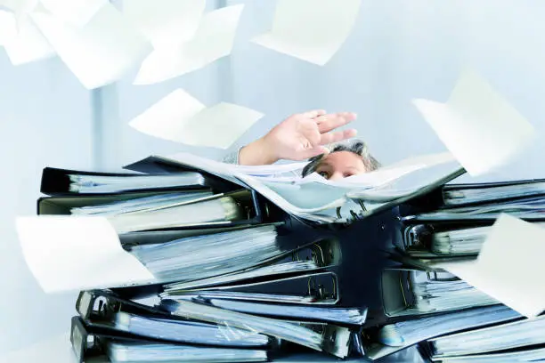 Photo of desperate woman behind high stacks of ring binders and lots of papers are flying around in the office, concept of excessive demands and increasing work in business