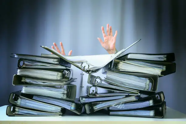 Photo of raised hands of a person who sinks behind stacks of ring binders on an office desk, concept of excessive demands and increasing work in business