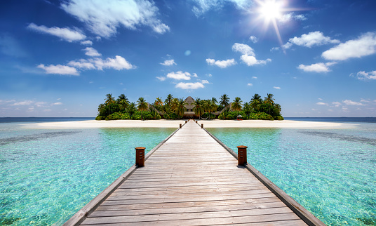 A wooden pier leading to a tropical paradise island with coconut palm trees, turquoise sea and sandy beaches
