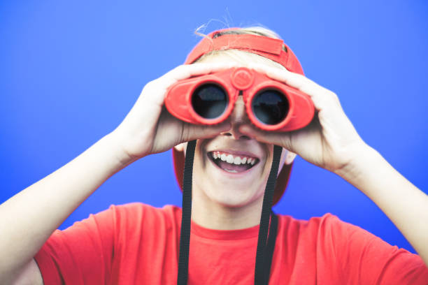 istock Beautiful boy looking away with a red binoculars isolated on a blue background. Colorful image of a young male having fun with a telescope. Fun, astonishment, happiness concept. Focus on the mouth. 1182110345