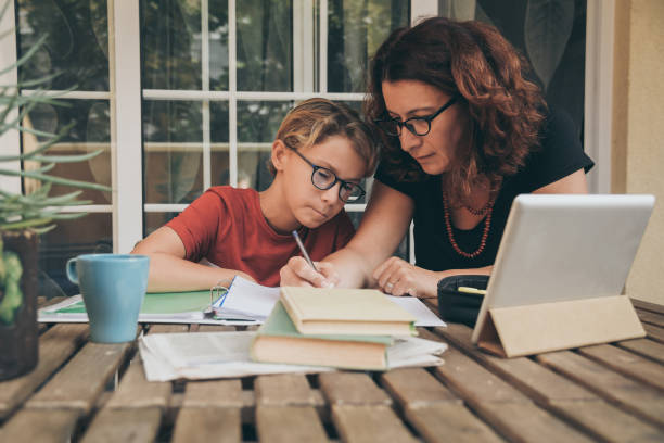 istock Young student doing homework at home with school books, newspaper and digital pad helped by his mother. Mum writing on the copybook teaching his son. Education, family lifestyle, homeschooling concept 1182111159