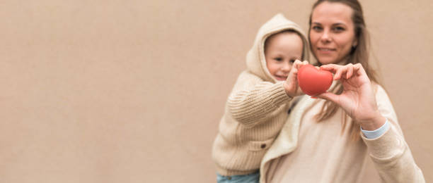 Mom woman with a little boy son 4-5 years old, background wall. In the hands of a toy red heart, concept give life and love, donation. Mother and St. Valentine's Day. Free space for text. stock photo