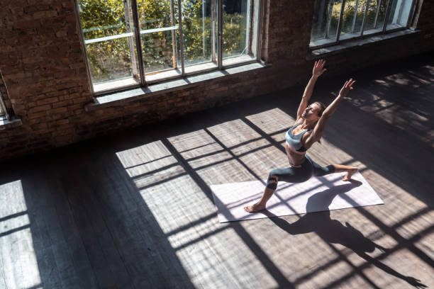 Sporty fit active yogi young woman in sportswear standing in warrior one pose on wooden floor mat in gym studio big windows, female healthy girl doing yoga exercise position workout concept, top view ajuste deportivo activo yogui joven mujer en ropa deportiva de pie en guerrero una pose en la alfombra de piso de madera en el estudio de gimnasio grandes ventanas, mujer chica sana haciendo concepto de entrenamiento de posición de ejercicio de yoga, vist