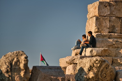 Amman, Jordan - October 15, 2017: Two men watching the sunset on the town ruins and enjoying their time in Amman, Jordan, with the national flag raised in the background.