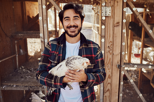 Portrait of a young farmer smiling while standing outside of a coop on his farm holding a chicken