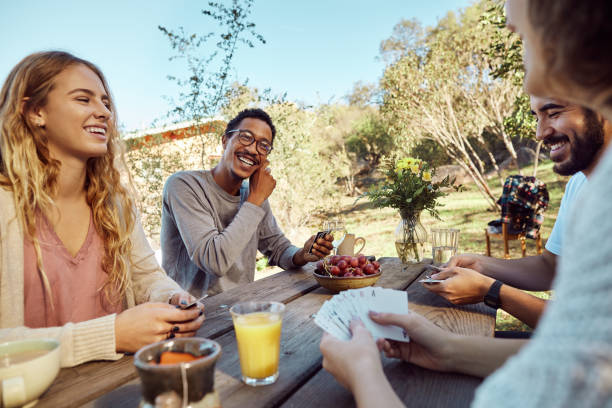 Laughing friends playing cards together outside at a picnic table Laughing group of diverse young friends playing cards together over breakfast at a picnic table outside friends playing cards stock pictures, royalty-free photos & images