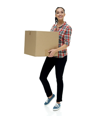 Full length / one person / side view / profile view of 18-19 years old adult beautiful black hair / long hair / ponytail caucasian female / young women standing in front of white background wearing jeans / pants / lumberjack shirt / plaid shirt / button down shirt / shirt / canvas shoe who is smiling / happy / cheerful / cool attitude who is picking up / packing / unpacking / moving house / relocation and holding box - container