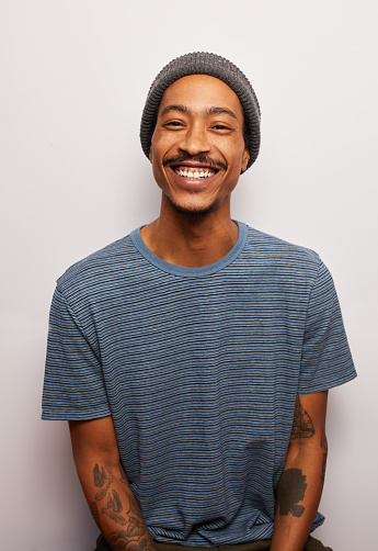 Studio portrait of a smiling young man with tattoos wearing a t-shirt and hat standing against a gray background