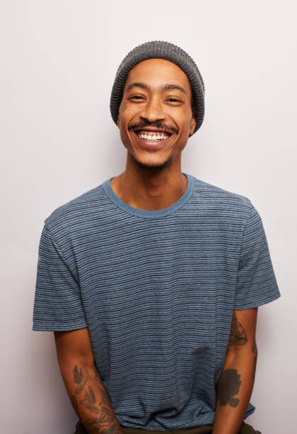 Smiling young man with tattoos standing against a gray background Studio portrait of a smiling young man with tattoos wearing a t-shirt and hat standing against a gray background alternative lifestyle stock pictures, royalty-free photos & images