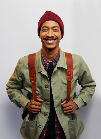 Studio portrait of a smiling young man ready for a hike with a jacket and backpack standing against a gray background
