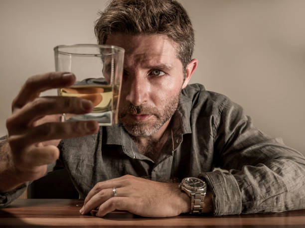 alcoholic depressed and wasted addict man sitting in front of whiskey glass trying holding on drinking in dramatic expression suffering alcoholism and alcohol addiction isolated on grey background stock photo