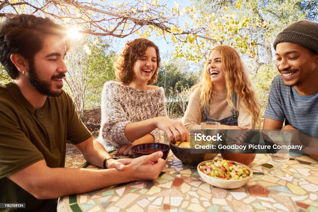 Laughing friends eating nachos togegher on an outdoor patio Group of diverse young friends laughing and eating snacks outdoors at a table on a farm Eating Stock Photo