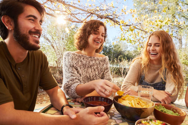 Laughing friends eating together outside on a sunny afternoon Group of young friends laughing and eating snacks together while sitting on a patio outdoors on a sunny afternoon tortilla chip photos stock pictures, royalty-free photos & images