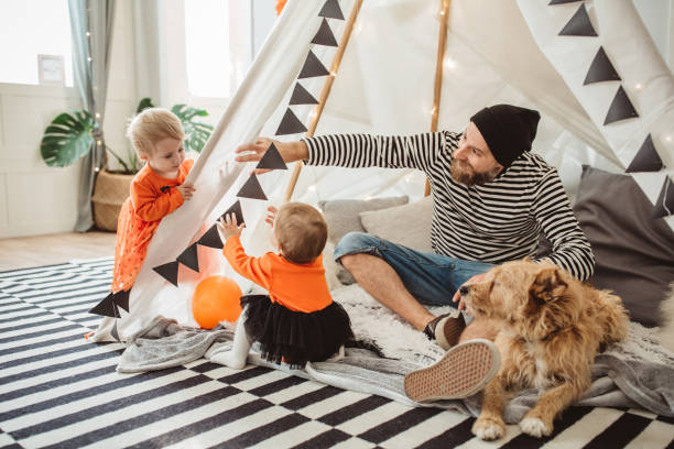 Playing in tent for Halloween Cheerful Father With daughters celebrating Halloween At Home. They fooling around and wear costumes, they sitting in tent, home is decorated, dog is with them too twin photos stock pictures, royalty-free photos & images