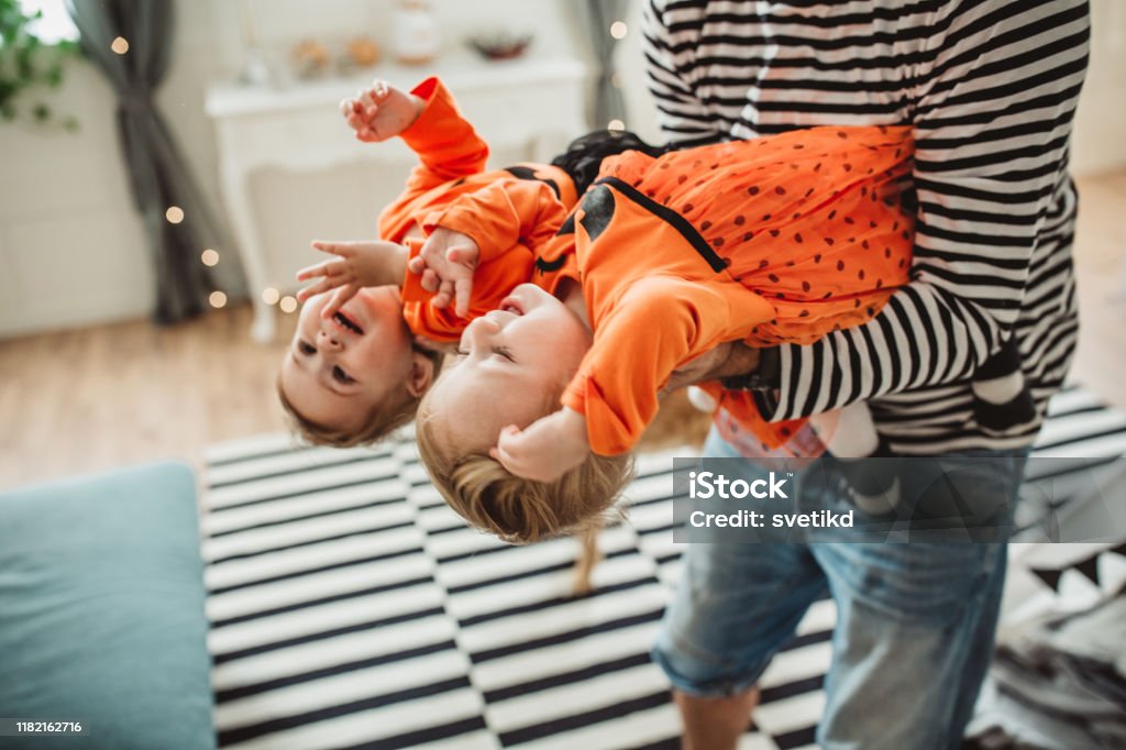 Happy family celebrating Halloween Cheerful Father With daughters celebrating Halloween At Home. They fooling around and wear costumes, father carrying them, home is decorated with flags Twin Stock Photo