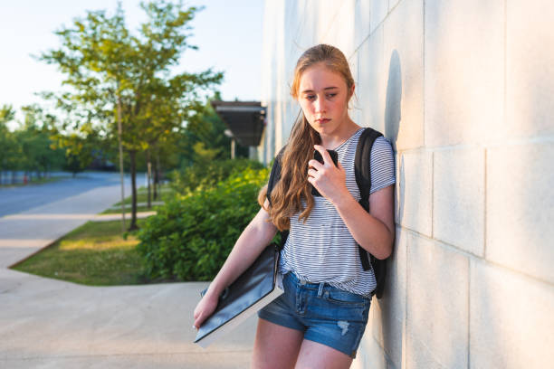 Depressed/Sad teen girl leaning against high school wall during sunset while wearing a backpack and holding binders/smartphone. stock photo