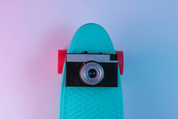 istock Plastic mini cruiser board and retro camera on background with blue pink neon gradient ligh. Top view 1182183068