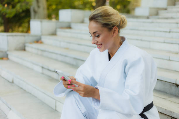 joven mujer atractiva en kimono blanco con cinturón negro. mujer deportiva sentada en las escaleras y utiliza el teléfono inteligente al aire libre. descansa después del entrenamiento. artes marciales - martial arts women tae kwon do black belt fotografías e imágenes de stock