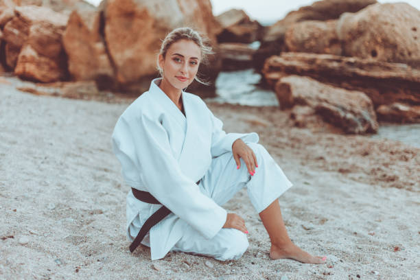 artiste martial féminin attirant dans le kimono blanc avec la ceinture noire se repose sur la plage sauvage de sable - martial arts women tae kwon do black belt photos et images de collection