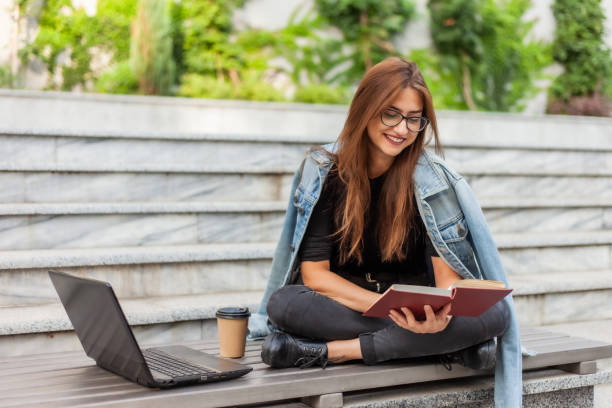 istock Modern students. Distance learning. Young enthusiastic woman reads book while sitting on bench with a laptop 1182194890