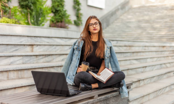 istock Modern students. Distance learning. Young hipster girl reads book while sitting on a bench with a laptop 1182194897