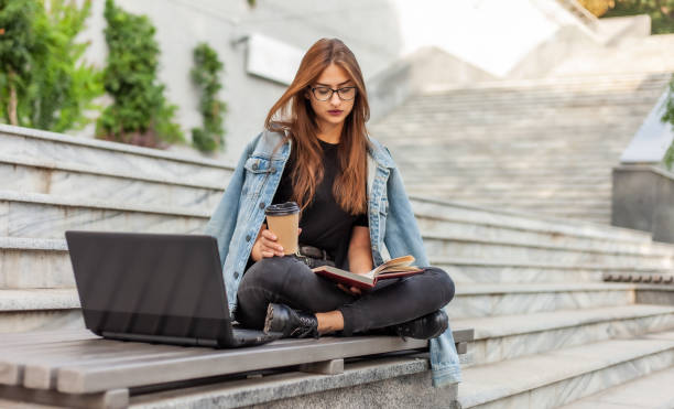 istock Modern students. Distance learning. Young hipster girl reads book while sitting on a bench with a laptop 1182194913