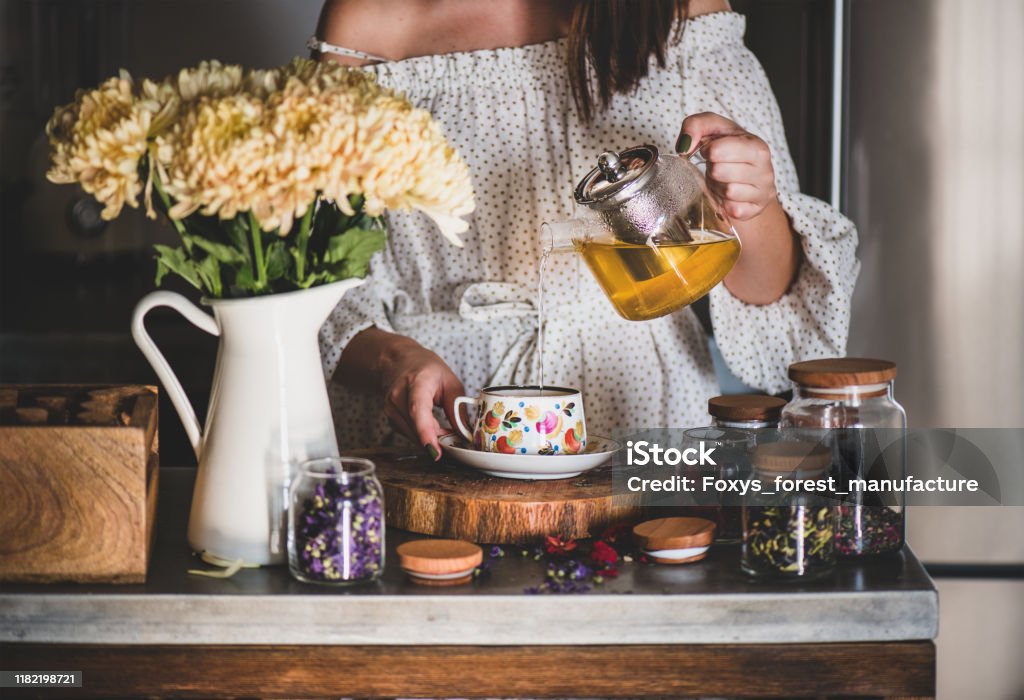 Young woman pouring green tea from pot into cup Young caucasian woman pouring freshly brewed green tea from glass pot into beautiful vintage porcelain cup at kithcen counter. Autumn tea time concept Tea - Hot Drink Stock Photo