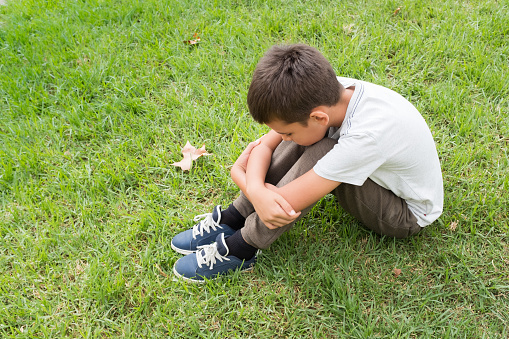 Side view of a child with brown hair wearing a short sleeved shirt and brown pants. He is looking down sitting on the grass at the park. Distraught, solitude and unhappiness concepts.