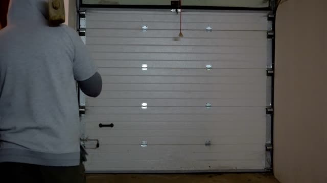 A man with a sledgehammer on his shoulder, in a hood, approaches the garage door