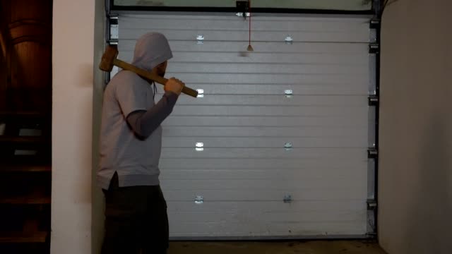 A man with a sledgehammer on his shoulder, in a hood, stands in the garage and looks around