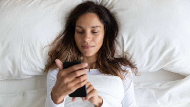 jeune fille mélangée paisible de course se réveillant vers le haut, utilisant le téléphone dans le lit. - hotel wireless technology bedroom hotel room photos et images de collection