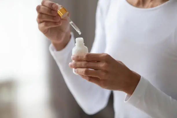 Close up young woman holding bottle with pipette, using coconut moisturizing oil or pleasant fragrance liquid. Lady opening container with medical cosmetic product, beauty procedure at home.