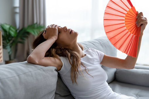 Side view exhausted young biracial girl using paper waver, suffering from hot summer weather or high temperature at home. Unhappy overheated millennial mixed race woman feeling uncomfortable indoors.