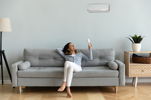 Front view full length joyful young mixed race woman relaxing on cozy couch in living room, holding remote controller, turning on cooler system air conditioner, setting comfortable temperature.