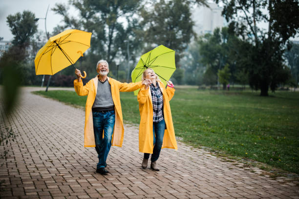 couples mûrs heureux dans les imperméables jaunes marchant avec des parapluies dans le stationnement. - umbrella senior adult couple autumn photos et images de collection