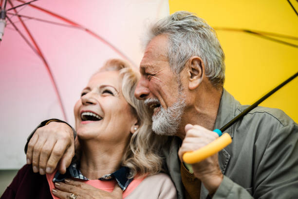 cheerful mature couple having fun while laughing under umbrellas. - umbrella senior adult couple autumn imagens e fotografias de stock