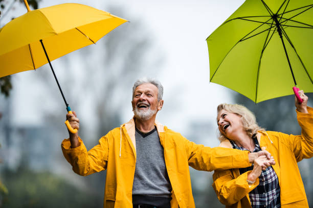 happy mature couple in yellow raincoats having fun with umbrellas in nature. - umbrella senior adult couple autumn imagens e fotografias de stock