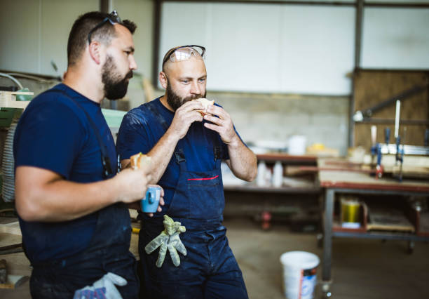 Young manual workers eating sandwiches on a break in a workshop. Young manual workers eating sandwiches on a lunch break in a workshop. construction lunch break stock pictures, royalty-free photos & images