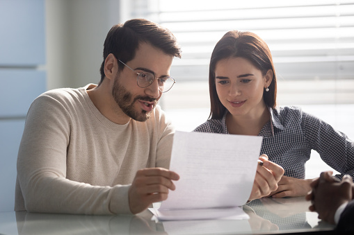 Young husband discussing contract details with smiling wife. Married couple reading carefully terms of conditions of paper document, making decision about house purchase or financial investment.