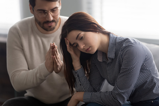 Head shot close up upset abused offended woman sitting on sofa, ignoring her apologizing husband. Young man feeling sorry, asking forgiveness to depressed wife. Family relationship problems concept.